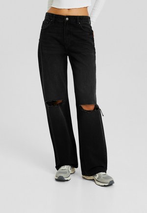 Femme portant un jean noir à jambes larges avec des genoux déchirés, un haut blanc crop top à manches longues, et des baskets grises, debout sur un fond uni.