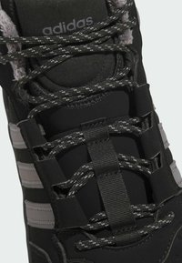 Zapatilla deportiva negra con una parte superior texturizada, franjas grises reflectantes y cordones moteados grises. Incluye ojales reforzados y un colchón acolchado.