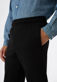 Hombre con pantalones de chándal negros con cintura elástica y camisa de botones de manga larga azul, con la mano en el bolsillo, logo de Champion visible en los pantalones.