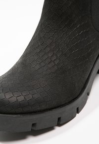Gros plan d'une botte noire avec un motif hexagonal texturé sur la tige et une semelle en caoutchouc épaisse et rainurée.