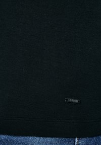 Tela negra con una pequeña etiqueta metálica rectangular que dice "G-STAR" cerca del dobladillo, combinada con jeans de mezclilla azul debajo.