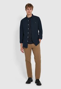 Camicia blu navy con bottoni bianchi, maniche lunghe. T-shirt nera sotto. Pantaloni slim-fit beige. Scarpe nere. Sfondo semplice.