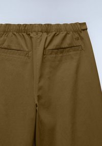 Braune elastische Bundhose, von hinten gezeigt, mit zwei horizontalen Schlitztaschen und Gürtelschlaufen auf einem schlichten hellen Hintergrund.