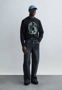 Μαύρο φούτερ με πράσινο γραφικό αστροναύτη, συνδυασμένο με άνετο σκούρο τζιν και μαύρα παπούτσια με κεντήματα λουλουδιών. Ανοιχτό μπλε καπέλο.