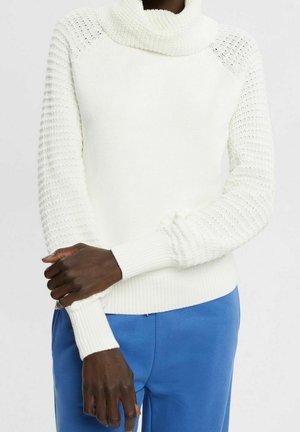 Personne portant un pull à col roulé en maille blanche texturée et un pantalon bleu, les bras croisés à la taille devant un fond uni.