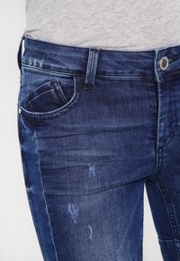 Tmavě modré denimmové džíny s hladkou texturou, které mají malou přední kapsu a jemné opotřebované detaily v oblasti stehna.