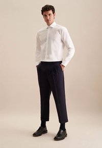 Camicia bianca con collo, pantaloni plissettati blu navy, scarpe nere e calzini. Tessuto liscio, vestibilità su misura e design della camicia corta.