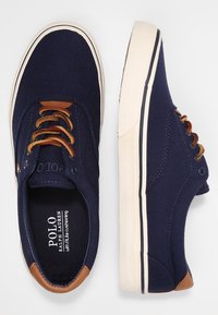 Námořnické plátěné tenisky s hnědými koženými akcenty, bílou podrážkou, kulatou špičkou a kontrastním prošitím. Disponují klasickým šněrovacím designem.