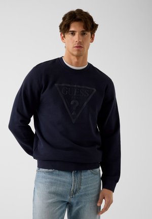 Jeune homme portant un sweat-shirt noir Guess et un jean bleu clair, debout devant un fond clair uni.