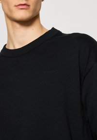 Gros plan d'une personne portant un t-shirt noir à col rond avec le logo "GANT" discrètement brodé sur la poitrine.