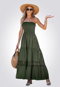 Robe maxi sans bretelles de couleur vert olive, avec un design à niveaux, un corsage smocké, des accents en dentelle et des poches. Portée avec un chapeau de paille et un sac.