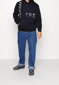 Black hoodie with white "TOM HILFIGER" branding, paired with blue denim jeans and grey sneakers. Hood and kangaroo pocket detail visible.