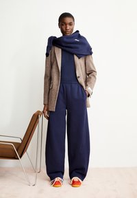 Marineblaue Jogginghose kombiniert mit einem braunen strukturierten Blazer und einem marineblauen Pullover, der über die Schultern drapiert ist; leuchtend rote Sneakers mit gelben Akzenten.