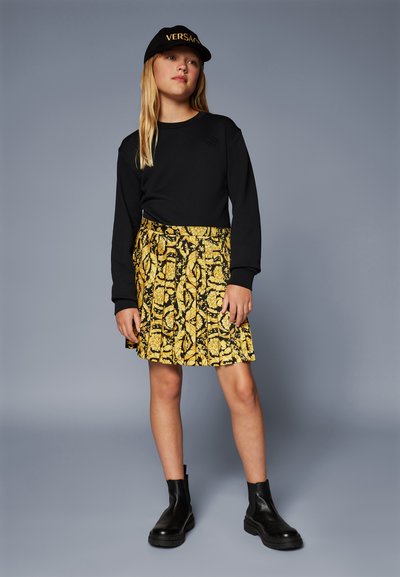 Top negro de manga larga, falda amarilla con tablas y patrón floral negro, y botines negros. Gorra negra con el logo dorado de "VERSACE".
