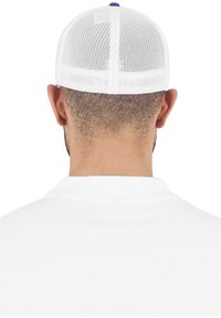 Vit mesh baseballkeps med en solid blå knapp på toppen; placerad bakom en man som bär en vit polo.