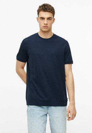 Jonge man met krullend haar die een marineblauw kortemouwenhemd en lichtblauwe spijkerbroek draagt, staat tegen een witte achtergrond.