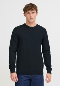 Giovane uomo con capelli ricci che indossa un maglione a maniche lunghe nero semplice e jeans blu scuro, in piedi contro uno sfondo bianco.