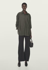 Camicia oversize verde scuro con bottoni, maniche arrotolate, abbinata a pantaloni neri a gamba larga e una piccola borsa texture.