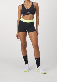 Schwarzer Sport-BH mit Ausschnitt, neon-gelbe Shorts mit Taillenbund, kombiniert mit schwarzen knielangen Socken und weißen Sportschuhen mit neonfarbenen Akzenten.