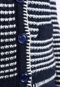 Cardigan lavorato a maglia blu navy con righe bianche, dotato di quattro bottoni blu e di una piccola tasca davanti, creando un motivo testurizzato.