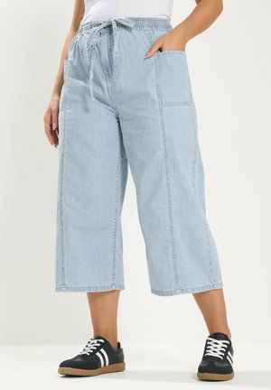 Person trägt hellblaue, weit geschnittene, kurz geschnittene Jeanshosen mit großen Seitentaschen und schwarze Turnschuhe mit weißen Streifen.