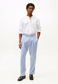 Pantaloni in lino azzurro chiaro con pieghe, tasche laterali e dettaglio con bottone, abbinati a una camicia bianca a maniche lunghe con bottoni. Mocassini marroni.