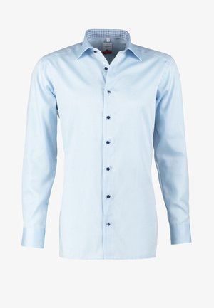 Uomo che cammina indossando una camicia azzurro chiaro con maniche arrotolate, pantaloni neri, scarpe marroni e portando una borsa a tracolla in pelle marrone.