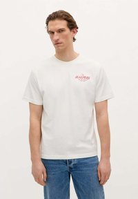 T-shirt en coton blanc à manches courtes et col ronde, avec un texte graphique rouge sur la zone gauche de la poitrine. Assorti à un jean en denim bleu.