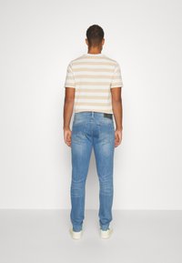 En man vänd bort från kameran iklädd en beige och vit randig t-shirt, ljusblå jeans och vita sneakers mot en helt vit bakgrund.