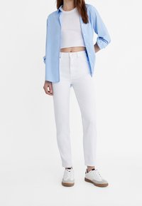 Světle modrá košile na knoflíky přes bílý crop top, kombinovaná s bílými džíny rovného střihu a bílými teniskami s gumovou podrážkou.