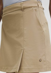Jupe beige longueur genou avec fente latérale, poche avant et petit logo rond près de l'ourlet portée avec un haut bleu clair.
