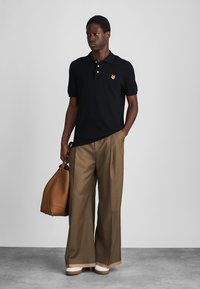 Must have särk musta polo, millel on väike tikitud rebane, koos heleda pruuniga laia säärega pükste ja pruuni nahkse kotiga. Valged spordijalatsid.