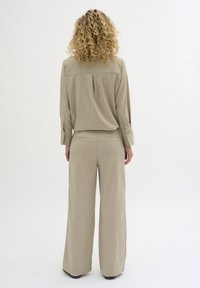 Béžový dvoudílný outfit obsahující košilový top s knoflíkovými detaily a kalhoty širokého střihu. Hladká textilie a volný střih, s tygřími sklady na zadní části topu.