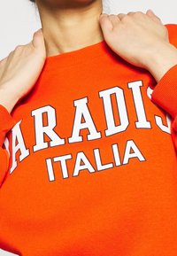 Oranžová mikina s výrazným bílým textem, který říká "PARADIS ITALIA." Žebrovaný límec a manžety s jemnou texturou.