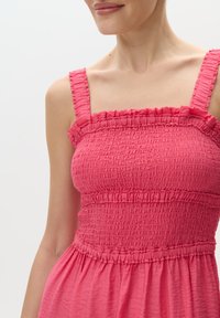 Różowa sukienka z falbanami, z grubymi ramiączkami; teksturowane, falujące detale przy dekolcie i w pasie, płynący materiał opadający poniżej talii.