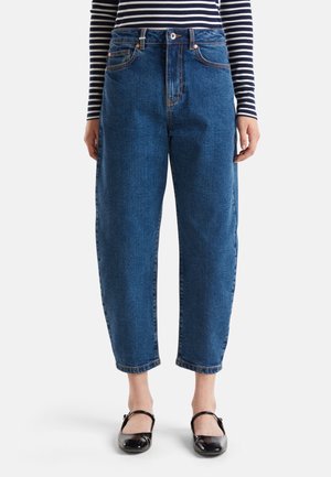 Vrouw draagt high-waist, loszittende blauwe jeans die boven de enkel zijn afgesneden, gecombineerd met zwarte Mary Jane schoenen en een gestreept longsleeve top.