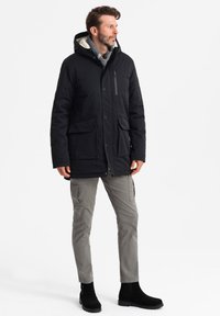 Parka nera con cappuccio e foderato in pile, dotata di due tasche frontali e una tasca con zip. Indossata con pantaloni cargo grigi e stivali neri alla caviglia.
