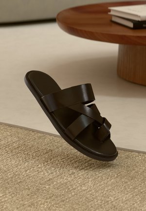 Sandale glissante en cuir noir avec trois sangles croisées, semelle plate et texture lisse, suspendue au-dessus d'un tapis en fibres naturelles tissées.