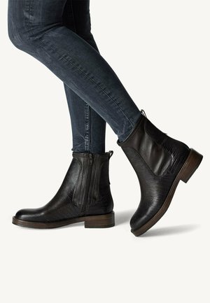 Botas de tobillo de cuero negro con acabado texturizado, cremallera lateral y tacón marrón. Combinadas con jeans ajustados de color azul oscuro.