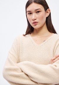 Béžový pletený sveter s výstrihom do V, s textúrovanou látkou a voľnými rukávmi. Jemný švový detail pozdĺž výstrihu.