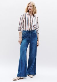 Camisa de botones a rayas con mangas largas, combinada con jeans de mezclilla azul de pierna ancha y alta cintura. Modelo lleva sandalias de tiras de color claro.