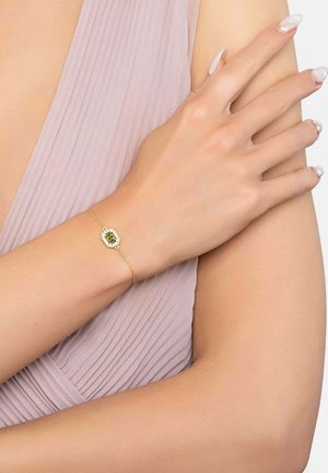 Finom arany karkötő zöld drágakővel és apró gyémántokkal a csuklón, halvány rózsaszín pliszírozott anyaggal és manikűrözött körmökkel párosítva.