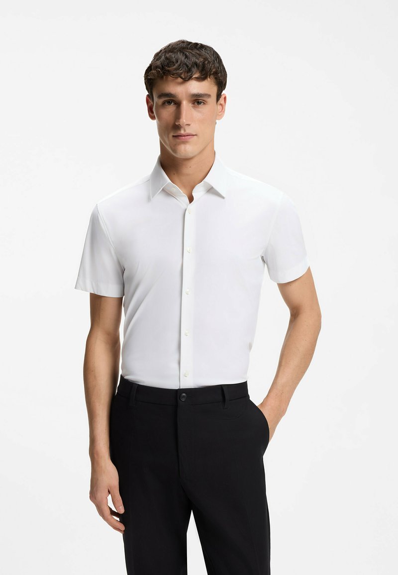 Homme portant une chemise blanche à manches courtes ajustée et un pantalon noir, debout avec une main dans la poche sur fond blanc.