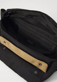 Black bag featuring a zippered main compartment, a tan fabric accent, and an internal zip pocket. Decorative hardware included.