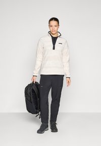Pullover in lichtroom met een patroonstructuur, zwarte kraag en knopen. Zwarte broek met ritszakken, zwarte rugzak en zwarte wandelschoenen.