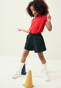 T-shirt rouge à manches courtes associé à un short noir à taille élastique. Chaussures de sport blanches et chaussettes blanches jusqu'aux genoux. Trois cônes de couleur à proximité.