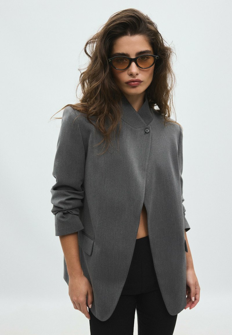 Femme aux longs cheveux ondulés portant des lunettes teintées marron, une veste grise boutonnée aux manches retroussées et un pantalon noir, debout devant un fond uni.