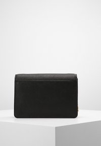 Schwarze strukturierte rechteckige Handtasche mit goldener Kettenriemen-Detail, auf einem weißen minimalistischen Podest vor einem schlichten weißen Hintergrund platziert.