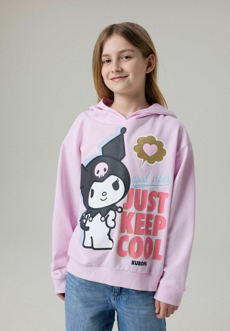 Jeune fille aux longs cheveux portant un sweat à capuche rose avec un personnage de dessin animé et le texte « Just Keep Cool » debout devant un fond uni.