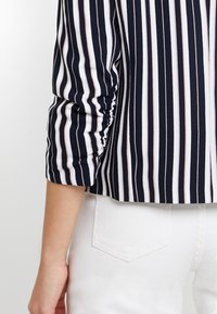 Haut rayé bleu marine et blanc à manches trois-quarts, avec un détail froncé à l'épaule. Le tissu semble lisse et extensible.